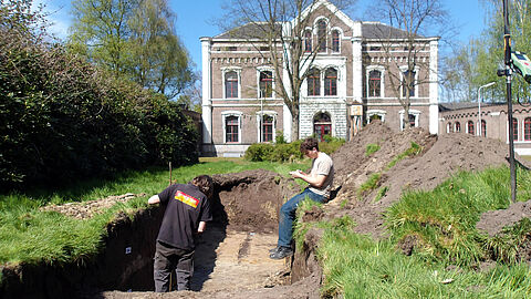 Twee archeologen staan en zitten in een gat in de grond, met op de achtergrond een landhuis.