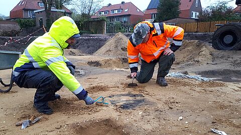 Twee archeologen met jassen aan bekijken een vondst in de grond.